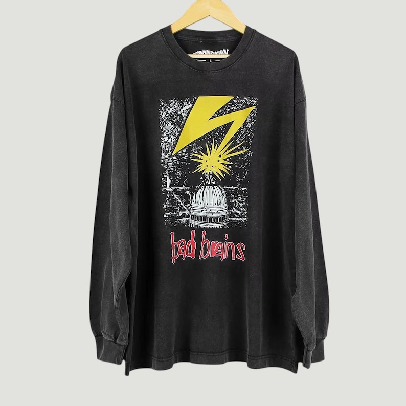 May include: A black long-sleeve t-shirt featuring a graphic print of a yellow lightning bolt striking a white building dome. Below the dome, the text "bad brains" is printed in red. The shirt has a crew neckline and a faded, washed-out appearance.