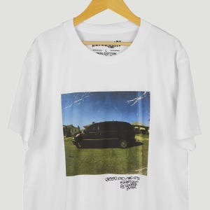 May include: White t-shirt with a graphic of a black van on grass under a blue sky. Text is at the bottom of the graphic. The shirt is made of 100% cotton.