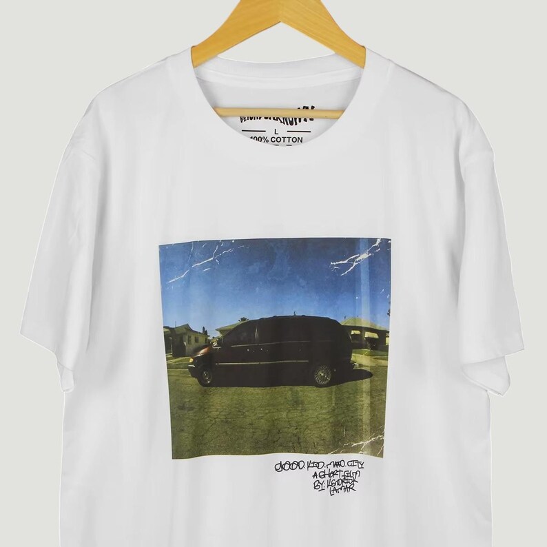 May include: White t-shirt with a graphic of a black van on grass under a blue sky. Text is at the bottom of the graphic. The shirt is made of 100% cotton.