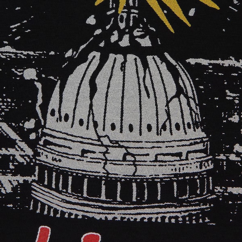 May include: A black fabric surface featuring a graphic print of a white domed building with visible cracks. Above the dome, yellow rays extend outward. Below the dome, partial red lettering is visible against the black background.
