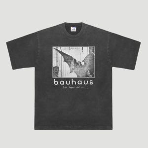 May include: Dark gray t-shirt featuring a black and white graphic with the band name "bauhaus" in white text. The graphic includes an image of a bat-like creature. The shirt has a crew neck and short sleeves.