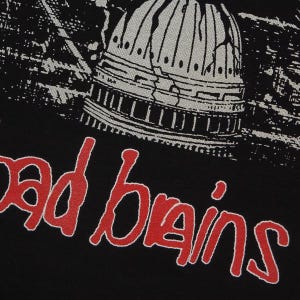 May include: A black t-shirt featuring a graphic print of a cracked, light gray dome structure above the red text "bad brains" in a stylized, hand-drawn font. The design is centered on the fabric, showcasing a high-contrast, detailed illustration.