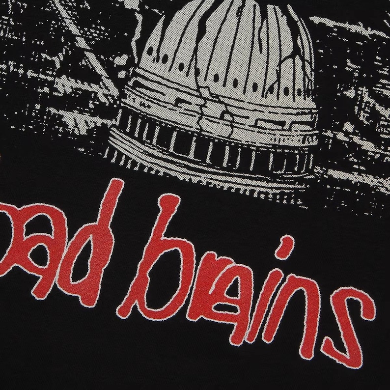May include: A black t-shirt featuring a graphic print of a cracked, light gray dome structure above the red text "bad brains" in a stylized, hand-drawn font. The design is centered on the fabric, showcasing a high-contrast, detailed illustration.