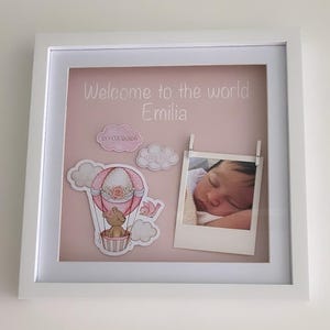 May include: A white framed baby announcement with a pink background. The text "Welcome to the world Emilia" is displayed. Decorative elements include a hot air balloon, clouds, and a photo of a sleeping baby. The date 20/03/2026 is also included.