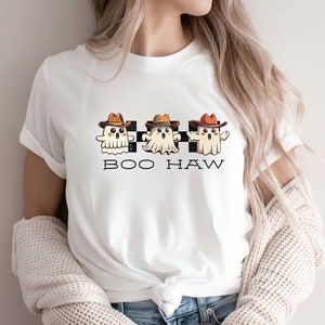 May include: White t-shirt featuring a design of three ghosts wearing cowboy hats and the text "BOO HAW" on a chequered background.