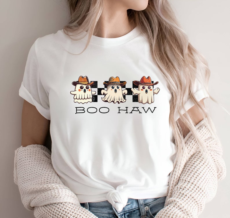 May include: White t-shirt featuring a design of three ghosts wearing cowboy hats and the text "BOO HAW" on a chequered background.