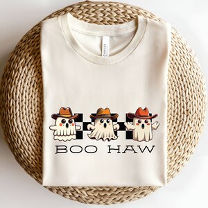 May include: A white t-shirt featuring a black and white chequered pattern with three cartoon ghosts wearing cowboy hats. The ghosts are white with orange and brown hats. The text "BOO HAW" is printed below the ghosts.