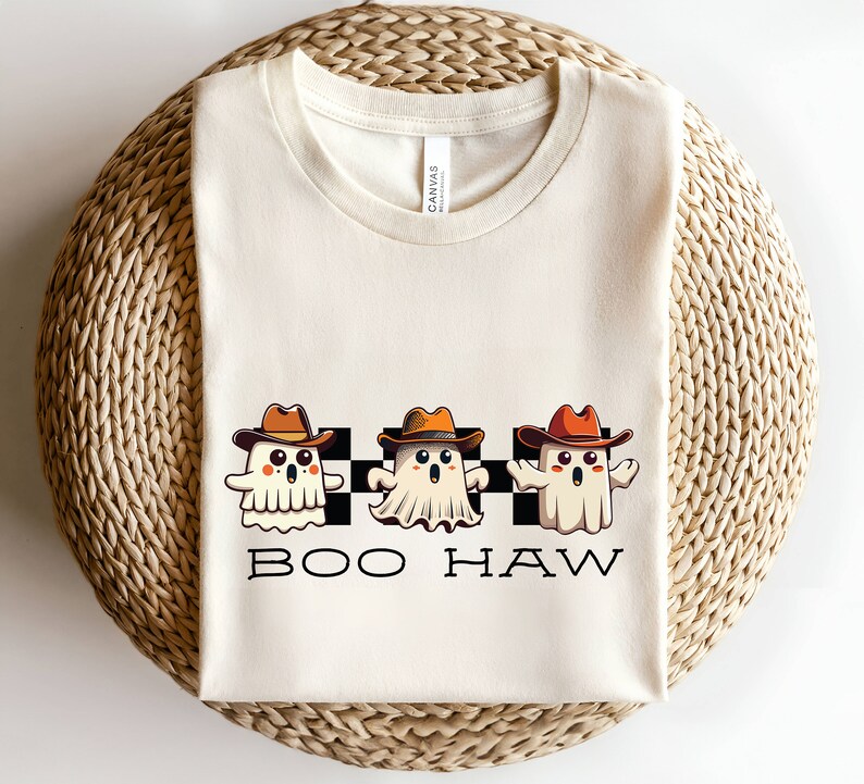 May include: A white t-shirt featuring a black and white chequered pattern with three cartoon ghosts wearing cowboy hats. The ghosts are white with orange and brown hats. The text "BOO HAW" is printed below the ghosts.