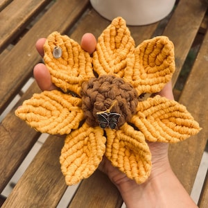 Può includere: Un girasole in macramè fatto a mano nei toni del giallo e del marrone. Il fiore presenta un'elaborata lavorazione dei petali e un centro strutturato. Piccoli ciondoli in metallo sono attaccati ai petali e al centro, aggiungendo un tocco di eleganza.