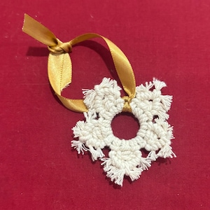 May include: A white, crocheted snowflake ornament with a gold ribbon for hanging. The snowflake has a circular center and fringed edges. The gold ribbon is tied in a knot. The ornament is set against a red background.