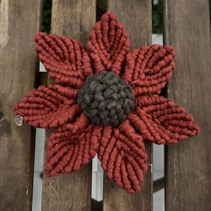 May include: A handcrafted macrame flower, featuring rust-coloured petals and a dark grey centre. The flower is displayed on a wooden surface. The intricate macrame pattern is clearly visible.