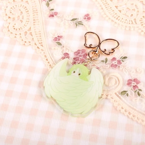 May include: A light green and white character with wings, printed on a clear acrylic charm. The charm is attached to a gold-colored metal chain with a decorative bow.