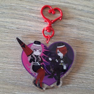 Puede incluir: Un llavero acrílico en forma de corazón con dos personajes de estilo anime vestidos de blanco y negro, sobre un fondo morado y rosa. Un cierre rojo en forma de corazón está adjunto.