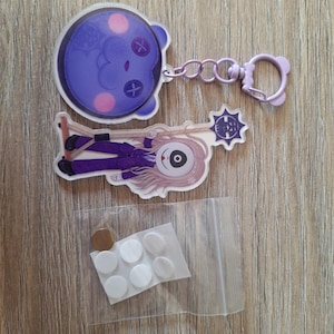 Può includere: Un portachiavi in acrilico a forma di orso viola con design a occhi a bottone, un secondo ciondolo in acrilico raffigurante un personaggio dai capelli lunghi con una grande maschera per gli occhi che tiene un bastone, e un sacchetto con adesivi bianchi.