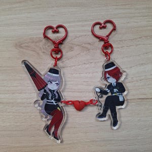 Puede incluir: Dos llaveros acrílicos con personajes de estilo anime. Un personaje sostiene un paraguas rojo y negro, el otro un cable. Ambos están conectados por un dije rojo en forma de corazón. Cada llavero tiene un cierre rojo en forma de corazón.