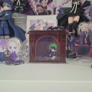 以下が含まれることがあります： アーチ型の開口部を持つダークレッドの暖炉フレームが特徴の長方形のアクリル製ディスプレイスタンド。フレームの内側には、緑色の髪と黒い眼帯をしたキャラクターの小さな平らなアクリル製ダイカットフィギュアが配置されています。