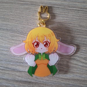 May include: A yellow and pink anime character keychain with bunny ears, wearing a green dress and a green bow. The keychain has a gold metal ring.
