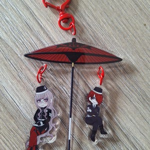 Könnte beinhalten: Ein roter herzförmiger Schlüsselanhänger mit einem roten und schwarzen Regenschirm-Anhänger. Der Regenschirm-Anhänger hat zwei transparente Acryl-Anhänger, die daran hängen, einer mit einer weiblichen Figur mit lila Haaren und der andere mit einer männlichen Figur mit roten Haaren.