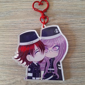 Könnte beinhalten: Ein Acryl-Schlüsselanhänger mit zwei Chibi-Anime-Figuren in dunklen Uniformen und Mützen. Eine Figur hat rotes Haar, die andere langes, hellviolettes Haar und eine Brille. Der Anhänger ist an einem roten, herzförmigen Metallverschluss befestigt.