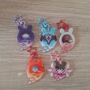 Puede incluir: Cinco llaveros acrílicos con personajes de dibujos animados con forma de mancha en varios colores. Cada llavero tiene un cierre en forma de estrella. Los personajes presentan diseños en morado, azul, rojo, naranja y rosa.