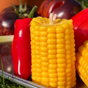 Può includere: Una candela a forma di pannocchia di mais gialla con uno stoppino bianco. La candela è circondata da peperoni rossi e una melanzana viola.