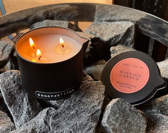 May include: A black candle jar with a white label that reads "coconut lime". The candle is lit and has three wicks. The jar is sitting on a bed of black rocks. The lid of the jar is on the rocks and has a pink label that reads "Massage Candle" with the text "Handmade With Love by CANDLE SWAN" underneath.