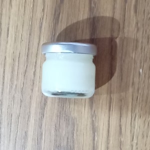 Puede incluir: Un pequeño frasco de vidrio transparente con tapa plateada, lleno de una sustancia blanca. El frasco está sobre una superficie de madera.