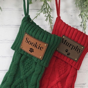 May include: Two personalized Christmas stockings, one green and one red, hang against a white brick wall. Each stocking has a brown leather patch with a name and paw print. The green stocking says "Sookie", and the red stocking says "Murphy".