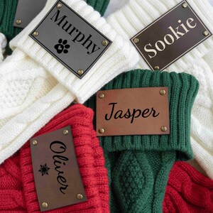 May include: Close-up of personalized Christmas stockings in red, white, and green. Each stocking has a leather tag with a name and decorative element, such as a paw print or snowflake. The stockings are made of a knitted material.