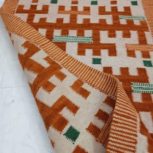 以下が含まれることがあります： 幾何学模様の折り畳まれた長方形のラグ。ラグは、オレンジと緑の形の繰り返し模様のあるクリーム色の背景が特徴です。端はオレンジとクリーム色のストライプで縁取られています。