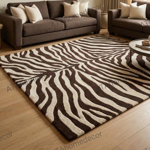 Op de afbeelding: Een rechthoekig vloerkleed met een bruin en crème zebrapatroon. Het kleed ligt op een lichte houten vloer voor twee donkerbruine banken met crèmekleurige sierkussens, een kamerplant en een kunstwerk aan de muur.