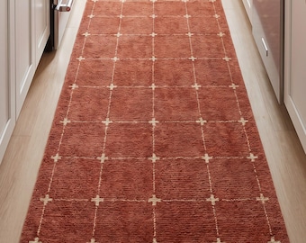 May include: A long, rectangular runner rug in a deep terracotta orange color featuring a white grid pattern with small cross accents at each intersection. The rug has a textured, pile surface and is positioned on a light-colored wooden floor.
