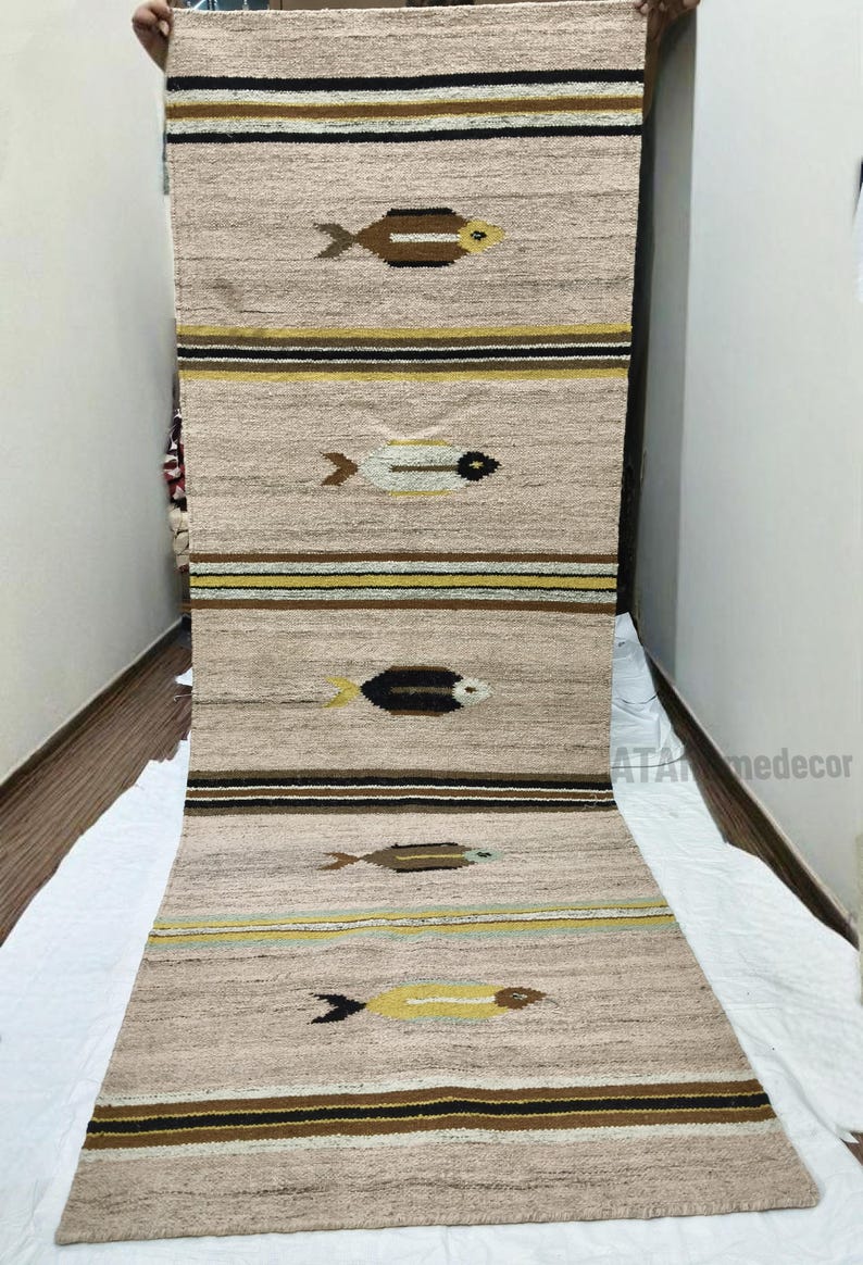 May include: Beige and brown woven rug with fish motifs. The rug features horizontal stripes in brown, yellow, and black. The fish are in various colors, including brown, yellow, and white. The rug is displayed vertically.