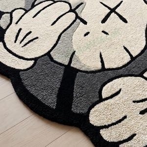 May include: A plush, tufted rug featuring a cartoon character's hands and face. The rug has a black border, gray and white sections, and black 'X' eyes. The hands are white with black outlines and details.