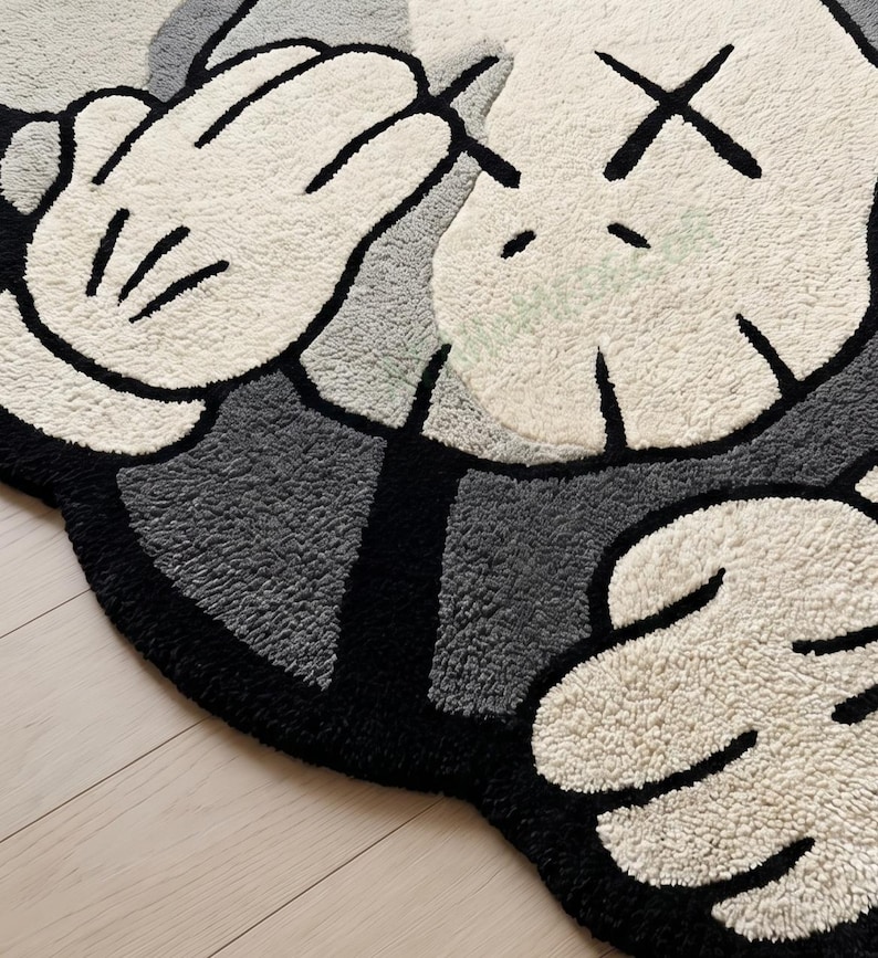 May include: A plush, tufted rug featuring a cartoon character's hands and face. The rug has a black border, gray and white sections, and black 'X' eyes. The hands are white with black outlines and details.