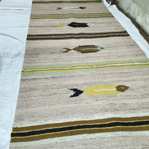 May include: A long, rectangular rug with a light beige background and horizontal stripes in brown, black, and yellow. The rug features repeating fish-like designs in brown, black, and white. The rug is made of a woven material.