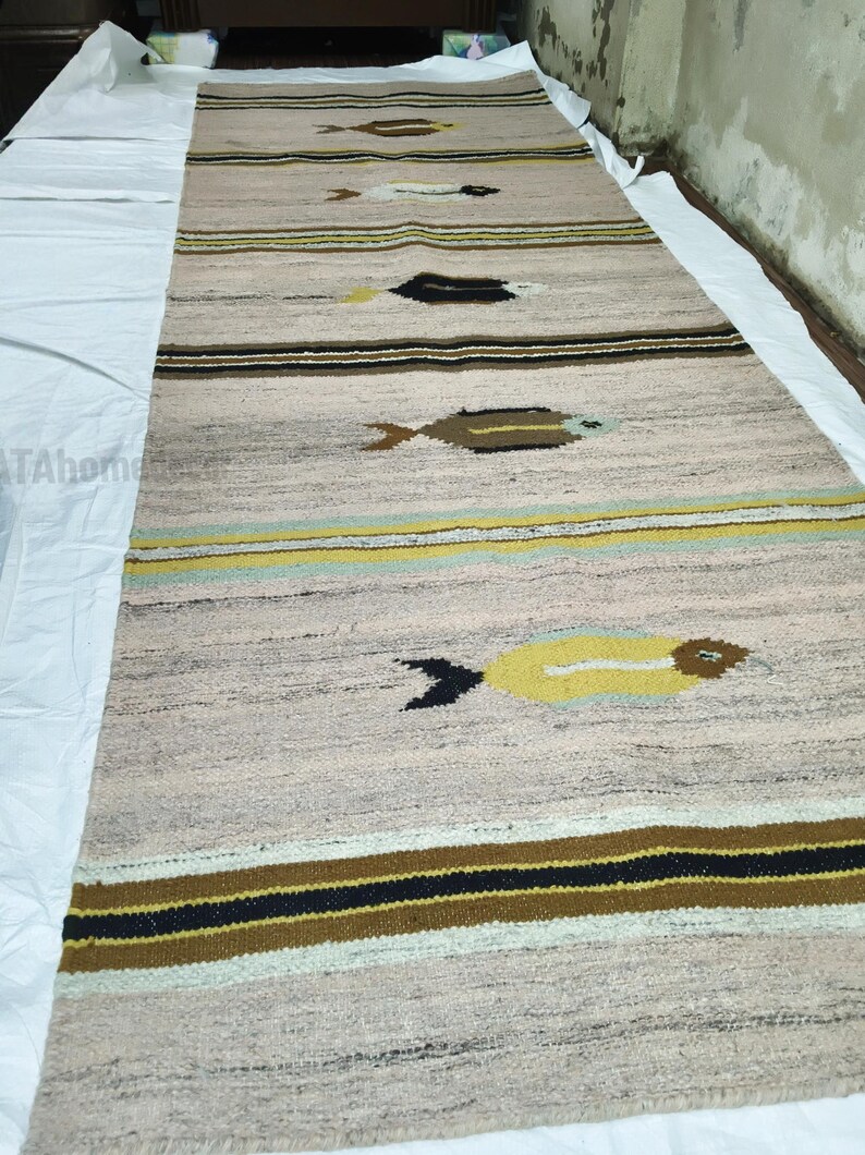 May include: A long, rectangular rug with a light beige background and horizontal stripes in brown, black, and yellow. The rug features repeating fish-like designs in brown, black, and white. The rug is made of a woven material.