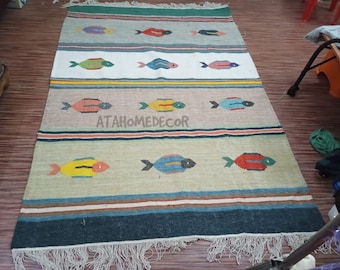 May include: Rectangular rug with a fish pattern. The rug features a beige background with rows of colorful fish in red, blue, yellow, and green. The rug has a fringe on the bottom edge and the text "ATAHOMEDECOR" in the center.