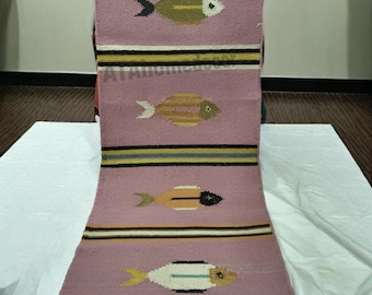 May include: A rectangular, hand-woven rug with a pink background and four fish motifs. Each fish is a different color, including gold, black, and white. The rug has horizontal stripes in black, yellow, and white.