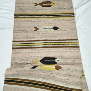 May include: A rectangular, woven rug with a light tan background. The rug features horizontal stripes in brown, black, and yellow. Three fish motifs in brown, yellow, and white are spaced along the rug's length. The rug is likely made of natural fibers.