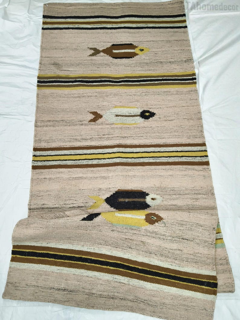 May include: A rectangular, woven rug with a light tan background. The rug features horizontal stripes in brown, black, and yellow. Three fish motifs in brown, yellow, and white are spaced along the rug's length. The rug is likely made of natural fibers.