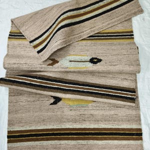 May include: Beige woven runner rug with a geometric pattern. The rug features stripes in black, brown, and gold. The design includes a central motif with arrow-like shapes. The rug is folded over itself.