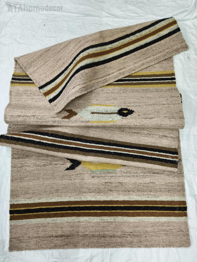 May include: Beige woven runner rug with a geometric pattern. The rug features stripes in black, brown, and gold. The design includes a central motif with arrow-like shapes. The rug is folded over itself.