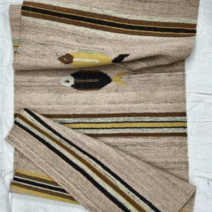 May include: A beige woven runner rug with a fish design. The rug features horizontal stripes in brown, black, and yellow. Two fish motifs are visible, one yellow and one black. The rug is made of a textured material.