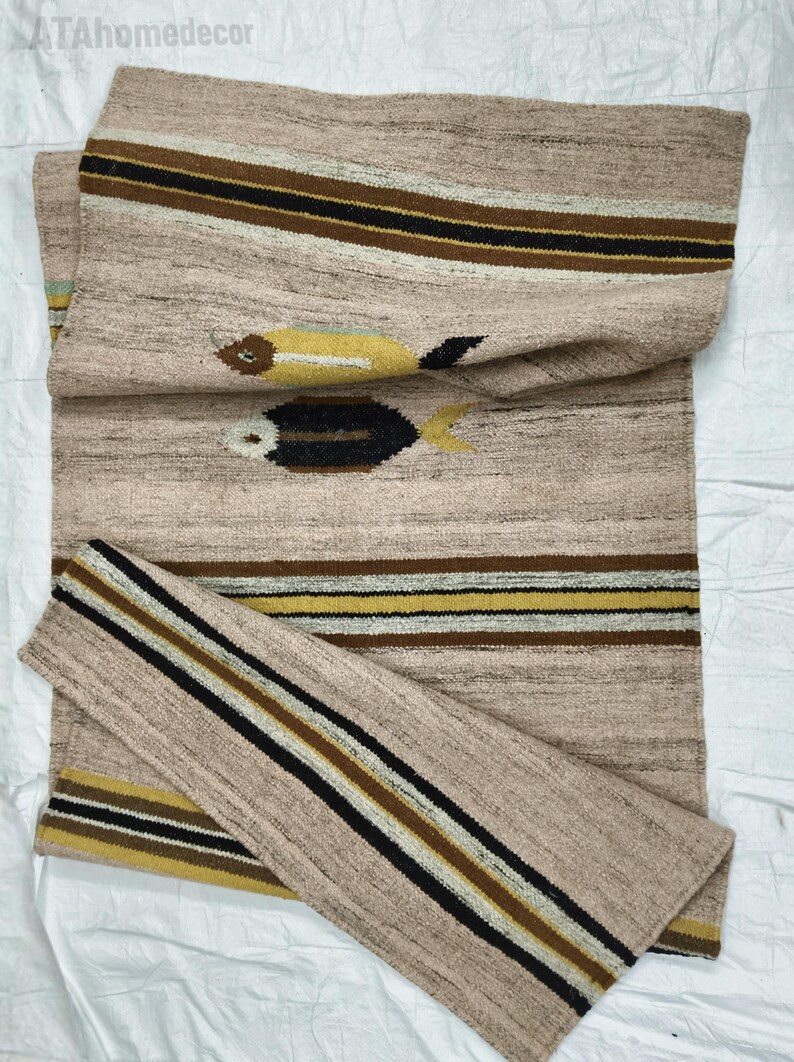 May include: A beige woven runner rug with a fish design. The rug features horizontal stripes in brown, black, and yellow. Two fish motifs are visible, one yellow and one black. The rug is made of a textured material.