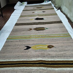 May include: A long, rectangular rug with a neutral beige background and horizontal stripes in brown, black, and yellow. The rug features repeating fish-like motifs in brown and yellow, creating a textured, patterned design.