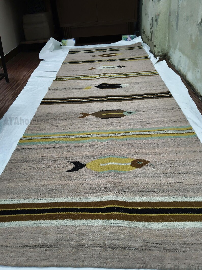 May include: A long, rectangular rug with a neutral beige background and horizontal stripes in brown, black, and yellow. The rug features repeating fish-like motifs in brown and yellow, creating a textured, patterned design.