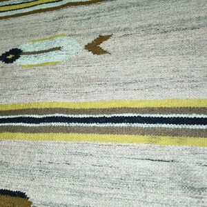 May include: A woven rug with a beige background and horizontal stripes in yellow, brown, and black. The rug features decorative feather-like and arrow-shaped designs in beige, brown, and black. The rug is made of a textured material.