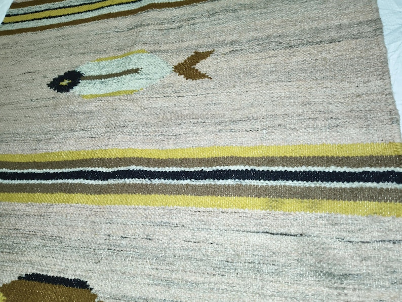 May include: A woven rug with a beige background and horizontal stripes in yellow, brown, and black. The rug features decorative feather-like and arrow-shaped designs in beige, brown, and black. The rug is made of a textured material.