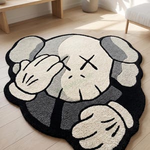 May include: A plush, character-shaped rug in shades of black, gray, and white. The rug features a cartoon-style design with a face and hands covering the eyes. The rug is placed on a light-colored wooden floor.